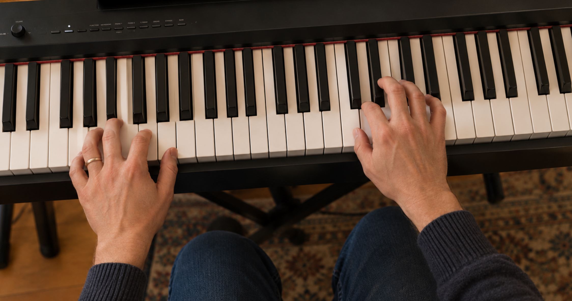 Overhead view of two adult hands on a digital piano keyboard, the right hand positioned higher playing a melody and the left hand lower holding a single bass note, both hands showing curved finger position, warm indoor light
