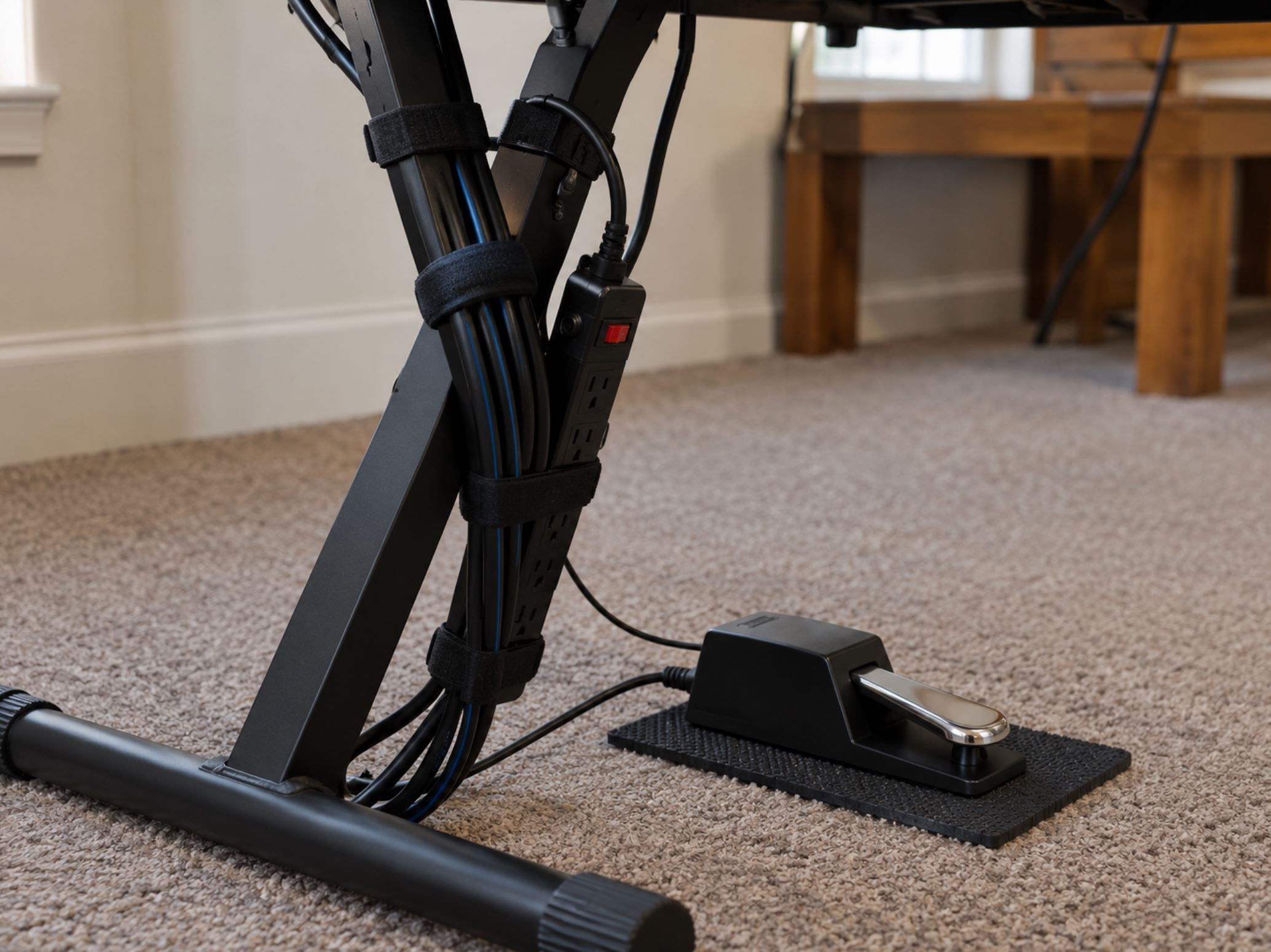 Close up of a keyboard stand leg with audio cables neatly secured using velcro ties, a power strip taped to the stand bar, a sustain pedal resting on a small rubber mat on carpet, practical and tidy