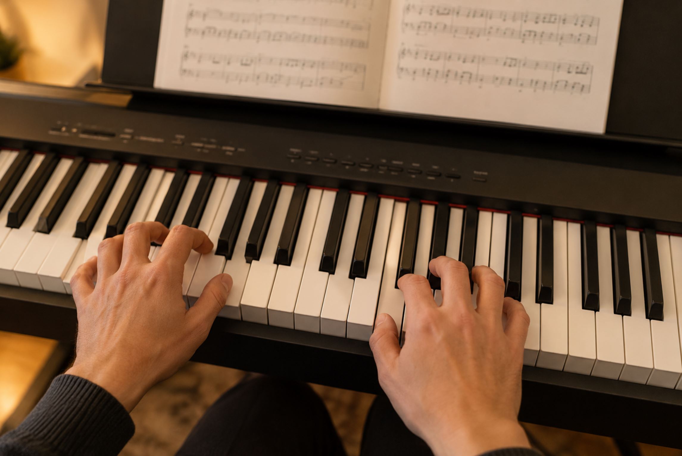 Close up of two adult hands playing an 88 key digital piano keyboard from above, fingers curved in proper position, open sheet music visible blurred in the background, warm indoor lighting