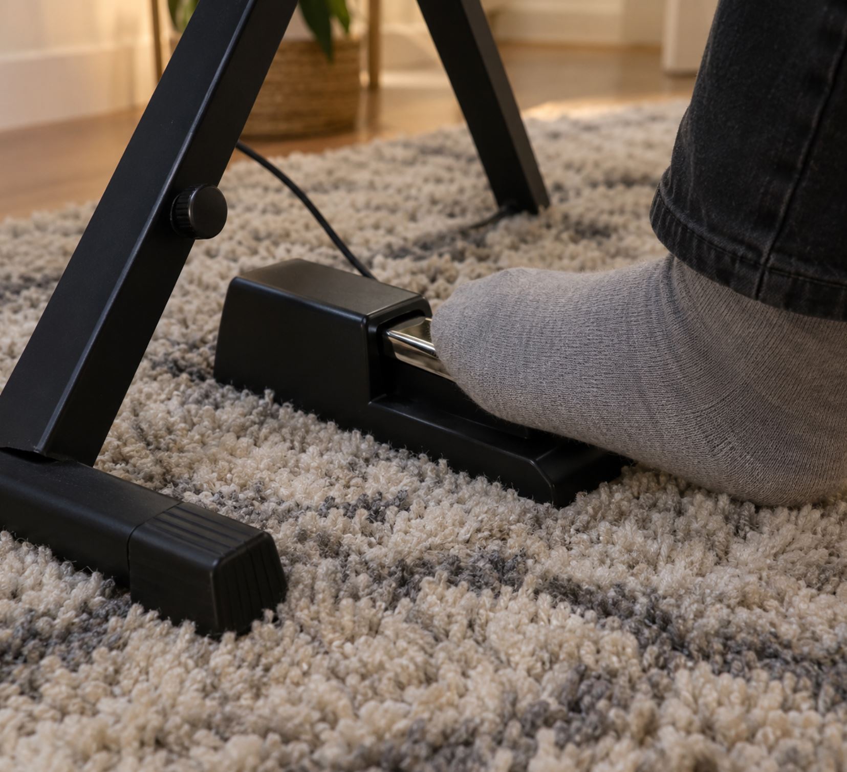 A sustain pedal resting on a thick rug beneath a keyboard stand, a foot in a sock resting on the pedal, close up view showing the rug fibres and the stand leg, soft domestic lighting