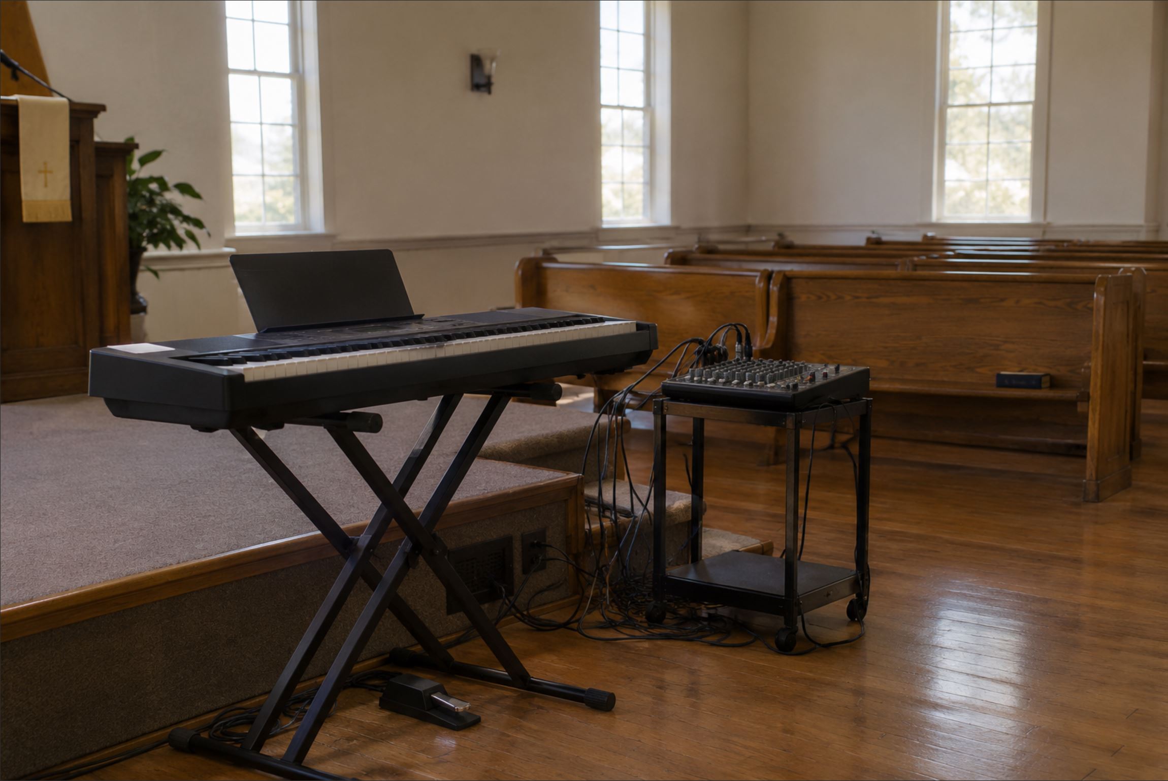 A keyboard on an X stand positioned to the side of a modest church platform, cables running to a small mixing desk, wooden pews visible in the background, natural light through tall plain windows, warm and unglamorous