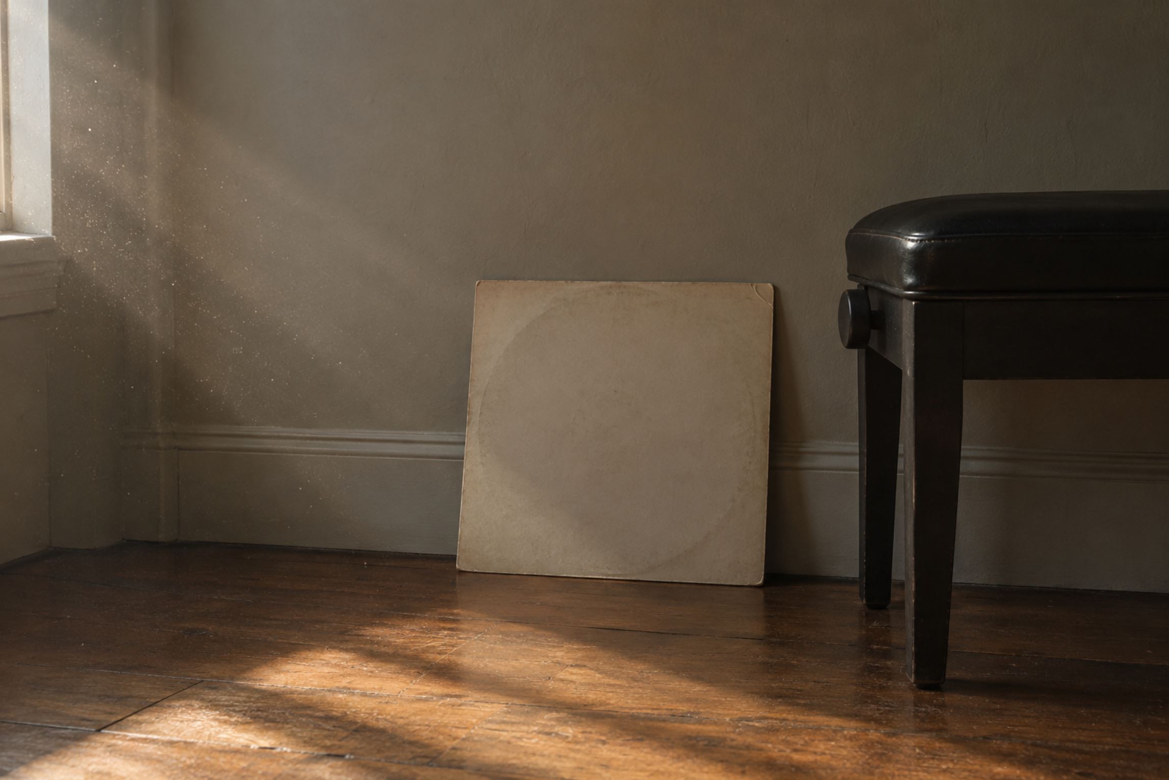 A plain record sleeve propped against a wall on a wooden floor beside a closed piano stool, soft side light, dust motes visible in the beam, muted colours, completely static composition